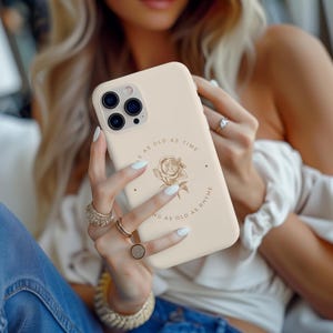 May include: A beige phone case with a gold floral design and the text "As old as time as old as rhyme".