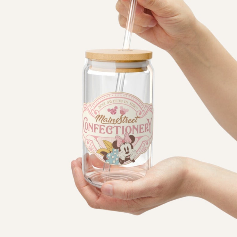 May include: A clear glass jar with a wooden lid and a straw. The jar has a pink and white label that says "Main Street Confectionery" with a cartoon image of Minnie Mouse. The label also says "Best Sweets in Town".