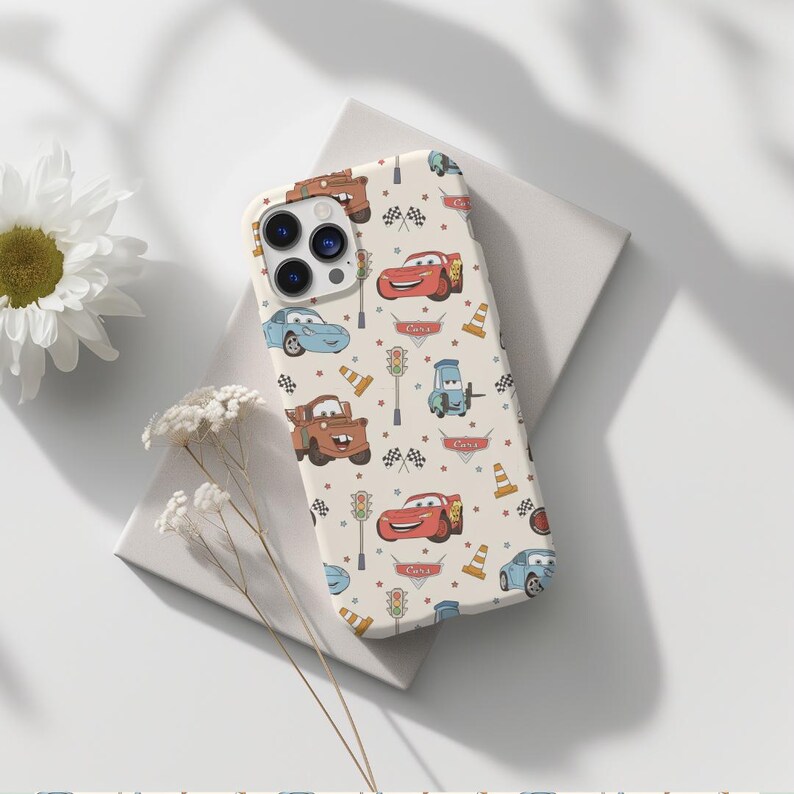 May include: A cream-colored smartphone case featuring a repeating pattern of characters and icons from the movie Cars, including red and blue race cars, brown tow trucks, traffic lights, orange traffic cones, and checkered racing flags.