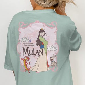 Może przedstawiać: Jasnozielony t-shirt w kolorze szałwii z grafiką na plecach przedstawiającą Mulan, Mushu i Little Brothera. Wzór zawiera ozdobną różową ramkę, stylizowane chmury oraz napis "LET YOUR INNER WARRIOR SHINE" nad imieniem "MULAN" ciemną czcionką.