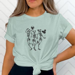 Puede incluir: Una camiseta de manga corta en color verde salvia claro con un estampado gráfico negro de tres esqueletos bailando. Cada esqueleto sostiene un globo negro con forma de cabeza de ratón. La camiseta tiene cuello redondo y está anudada en la cintura.