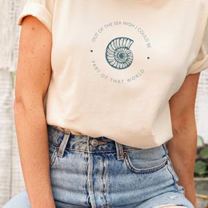 May include: A light beige t-shirt with a graphic of a seashell and the text "Out of the sea wish I could be part of that world".