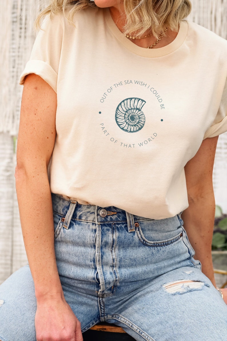 May include: A light beige t-shirt with a graphic of a seashell and the text "Out of the sea wish I could be part of that world".