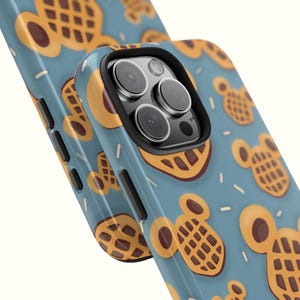 May include: A blue phone case featuring a repeating pattern of cartoon waffles in shades of brown and yellow. The case has a black camera lens border and a glossy finish. Sprinkles are scattered across the background.