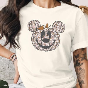 Puede incluir: Una camiseta de manga corta de color crema con un gráfico central de una calabaza con forma de cabeza de ratón. La calabaza tiene un estampado floral en tonos rosa, marrón y naranja, con un contorno negro que forma una cara sonriente y orejas.