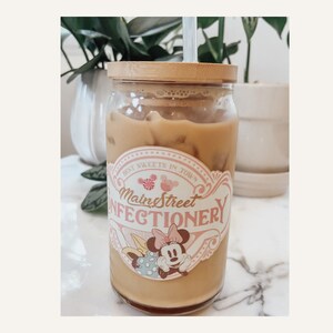 May include: A glass jar with a bamboo lid filled with iced coffee. The jar has a sticker with the text "Main Street Confectionery" and a cartoon image of Minnie Mouse.