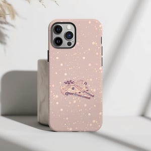 Puede incluir: Una funda de teléfono rosa con una ilustración detallada de la nave espacial Millennium Falcon. La funda tiene un patrón moteado y un módulo de cámara negro. El diseño es un guiño a la ciencia ficción.
