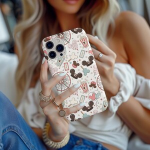 May include: A smartphone case featuring a repeating pattern of amusement park icons including ferris wheels, popcorn buckets, tickets, balloons, and mouse-ear silhouettes in shades of brown, cream, red, and light blue on a cream background.