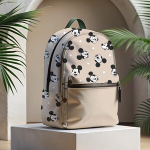 Puede incluir: Una mochila beige con una cremallera verde y un patr&oacute;n repetido de cabezas de Mickey Mouse sobre un fondo blanco.