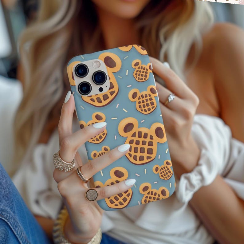 May include: A light blue phone case featuring a pattern of cartoon waffle designs in shades of brown and yellow. The case has a camera cutout and is held by a person with multiple rings.