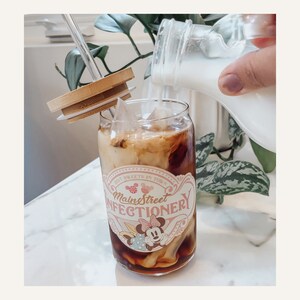 May include: A glass with a straw and a lid, filled with iced coffee and milk. The glass has a pink and white design with the text "Main Street Confectionery" and a cartoon image of Minnie Mouse.
