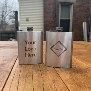 May include: Two silver hip flasks with engraved designs. One flask has the text "Your Logo Here" and the other has a diamond shape with the text "Smith" inside.