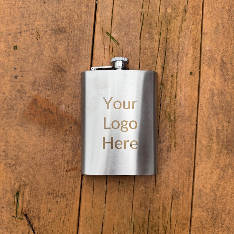 May include: A silver flask with a hinged lid and a cap. The flask has the text "Your Logo Here" engraved on the front.