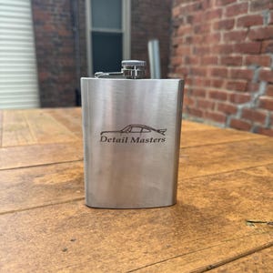 May include: A silver flask with a black and white car silhouette and the text "Detail Masters" engraved on the front.