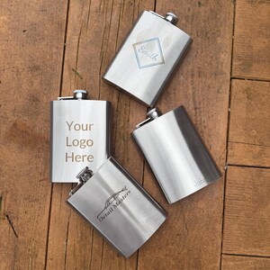 May include: Four stainless steel hip flasks with different engravings. The flasks are silver in color and have a rounded rectangular shape. The engravings include "Smith", "Your Logo Here", "Detail Masters", and "John".