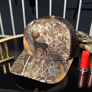 May include: Camouflage baseball cap with a brown mesh back and a deer silhouette patch. The cap features a Realtree camo pattern and a metal button on the side. A hunting-themed accessory.