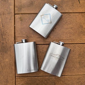 May include: Three silver hip flasks with personalized engravings. The flasks are on a wooden surface. The engravings include "Smith", "Johnson", and "Detail Masters".