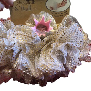 May include: A gold and white tulle tutu with gold glitter dots. A pink crown with gold glitter accents sits on top of the tulle. The tutu is for a baby and is labeled "Fancy Tutu Set - Size: 0-6M".