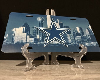 May include: A blue and white Dallas Cowboys license plate featuring a skyline of Dallas, Texas with the Dallas Cowboys star logo in the center.
