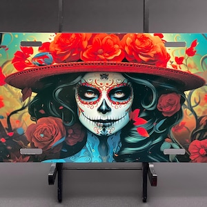 May include: License plate featuring a Day of the Dead design. The artwork depicts a woman with sugar skull makeup, wearing a red hat adorned with roses. The background is filled with flowers and foliage.