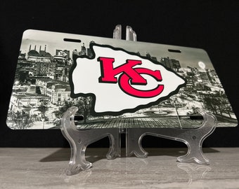 May include: A black and white image of a city skyline with a red and white "KC" logo on a white arrowhead. The image is on a metal license plate with a clear plastic stand.
