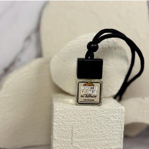 May include: A black and white car air freshener with a black string. The air freshener is a small bottle with a black cap and the text "MOROCCAN CASHMERE oil diffuser" printed on the front.