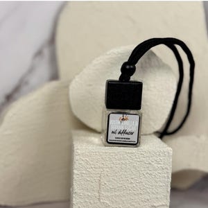 May include: A black and white car air freshener with a black string. The air freshener is a small glass bottle with a black cap and a label that reads "Warm Vanilla & Brown Sugar Oil Diffuser".