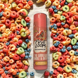 Può includere: Un tubo di balsamo per labbra rosa con le parole "Fruit Loops" e "For Chapped Lips" stampate su di esso. Il tubo è circondato da pezzi di cereali colorati, tra cui rosso, giallo, arancione e blu.