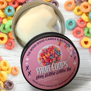 Puede incluir: Una lata rosa y blanca con una ilustración colorida de cereal. El texto en la lata dice "Fruit Loops Body Butter Lotion Tin" y "Shea, Mango & Cocoa Butters Almond Oil & Vitamin E". La lata es de 1,5 oz y hecha a mano en Atlanta, Georgia.