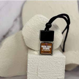 May include: A black and clear glass car air freshener with a black string hanging from it. The air freshener is labeled "Warm Fudge Brownie Oil Diffuser".
