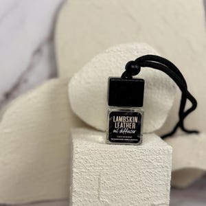 Puede incluir: Ambientador de coche negro con una cuerda negra colgando de él. El ambientador es una pequeña botella de vidrio con una etiqueta negra que dice "Lambskin Leather oil diffuser".