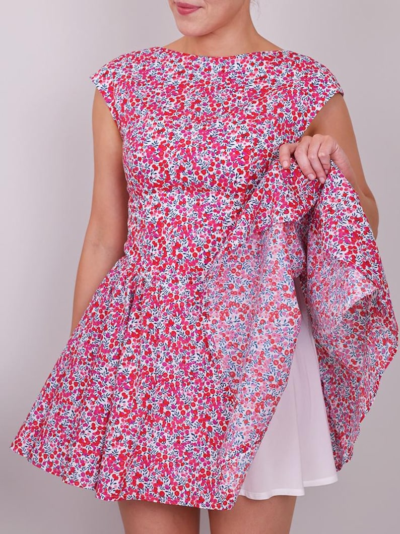 Puede incluir: Un vestido sin mangas con un estampado floral en tonos rojos y rosas sobre un fondo blanco. El vestido tiene un cuello barco y una falda acampanada. Se ve un enagua blanca debajo.