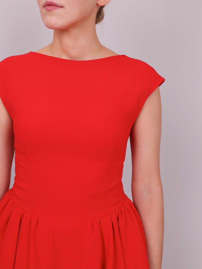 Puede incluir: Un vestido rojo vibrante con cuello barco y mangas cortas. El vestido presenta un corpi&ntilde;o ajustado que se ensancha en una falda plisada. El color brillante y el dise&ntilde;o cl&aacute;sico crean un look audaz y elegante.