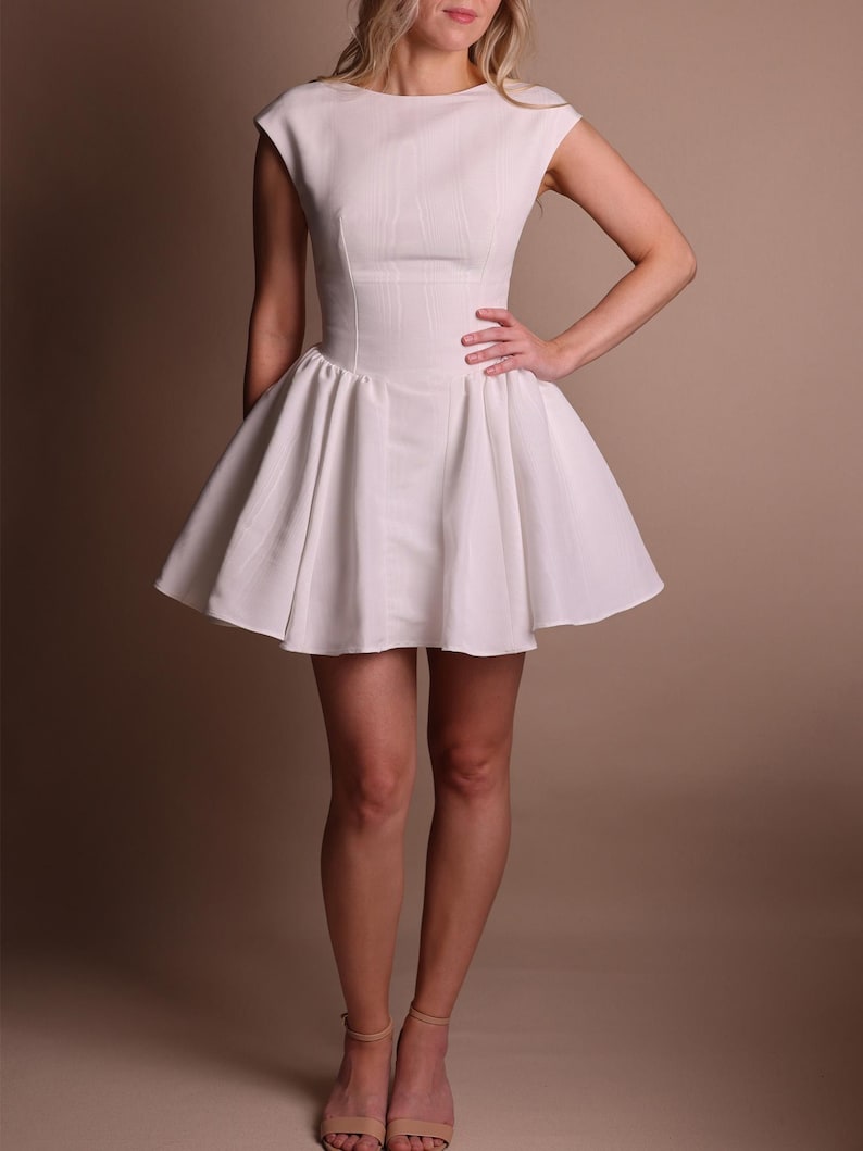 Puede incluir: Un vestido blanco con un corpi&ntilde;o ajustado y una falda acampanada. El vestido presenta un cuello redondo y mangas cortas. La modelo lleva tacones color nude. El fondo es de un color neutro s&oacute;lido.