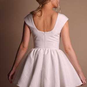Puede incluir: Un vestido blanco con espalda cuadrada y mangas cortas. El vestido presenta un corpi&ntilde;o ajustado y una falda acampanada. El modelo se ve desde atr&aacute;s, con los brazos ligeramente extendidos. El vestido est&aacute; hecho de una tela lisa y opaca.