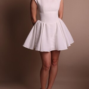 Puede incluir: Un vestido blanco con un corpi&ntilde;o ajustado y una falda acampanada. El vestido tiene un escote redondo y mangas cortas. La modelo lleva tacones color nude. El vestido est&aacute; hecho de una tela lisa y opaca.