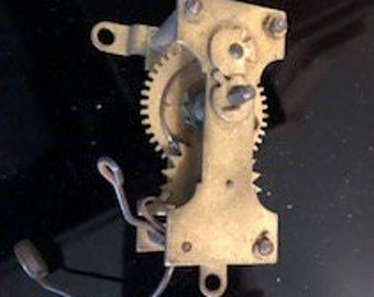 May include: A brass clock mechanism with gears and a winding key. The mechanism is mounted on a metal plate with four screws.
