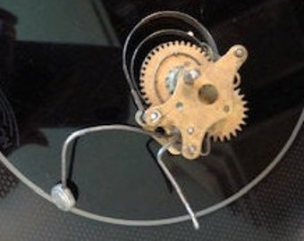 May include: Close-up of a vintage record player mechanism with brass gears and a metal arm. The mechanism is mounted on a black surface.