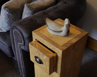 May include: A wooden side table with a drawer and a decorative duck figurine on top. The table has a light brown finish and a distressed look. The drawer has a metal handle.
