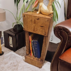 Op de afbeelding: Een houten bijzettafel met een plank voor boeken. De tafel is gemaakt van gerecycled hout en heeft een rustieke afwerking. Twee boeken staan op de plank, een met een kleurrijke kaft en de andere met een blauwe kaft. Twee kleine, gouden konijnenbeeldjes staan op de tafel.