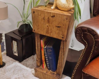 May include: A wooden side table with a shelf for books. The table is made of reclaimed wood and has a rustic finish. Two books are on the shelf, one with a colorful cover and the other with a blue cover. Two small, golden bunny figurines are on top of the table.