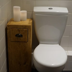May include: A white toilet with a wooden cabinet with a drawer underneath. Two white candles are on top of the cabinet.