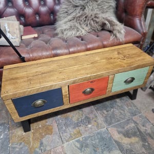 May include: A rustic wooden coffee table with three drawers. The drawers are painted navy blue, orange, and mint green, each with a brass-colored pull. The table has a natural wood top and black legs. This piece is ideal for living room decor.