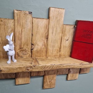 May include: A rustic wooden shelf with a natural wood finish. The shelf is made of reclaimed wood and has a unique, handcrafted look. A small gray rabbit figurine is on the shelf, and a red book with the text "Book of Big Plans" is on the right side of the shelf.