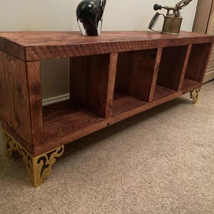 Peut inclure: Une étagère en bois avec quatre compartiments et des pieds décoratifs en métal doré. L'étagère est teintée d'une couleur brun foncé.
