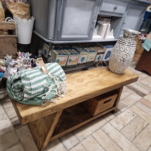 Op de afbeelding: Een rustieke houten bank met een driehoekige plank en een klein houten doosje met het opschrift "Gift". De bank is gemaakt van natuurlijk hout met een donkerbruine vlek. Een groen-wit gestreepte handdoek ligt op de bank.