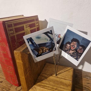 May include: A wooden photo holder with three metal clips holding up three instant photos. The photos show a dog in a car, a group of three people, and a close-up of one of the people. The holder is sitting on a wooden surface.