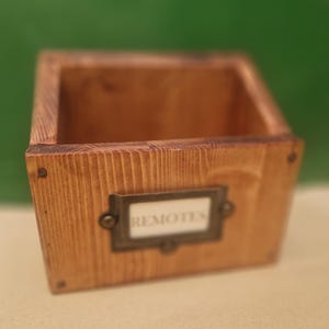 Op de afbeelding: Een rechthoekige houten opbergdoos met een natuurlijke bruine houtnerfafwerking. De voorzijde is voorzien van een donkere metalen labelhouder met een wit inzetstuk met de zwarte tekst "REMOTES". De doos is geconstrueerd met zichtbare metalen bevestigingen.