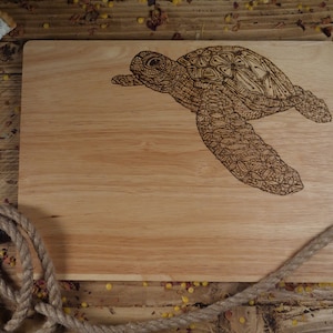 May include: A wooden cutting board with a detailed, intricate design of a sea turtle burned into the surface. The board is light brown and has a natural wood grain.