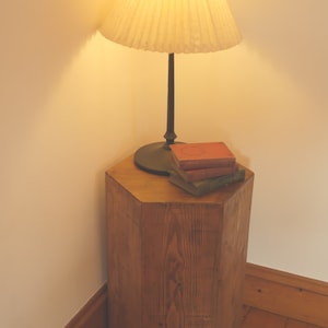 May include: A wooden hexagonal side table with a lamp and books on top. The lamp has a pleated white shade and a dark brown base. The books are red, green, and brown.